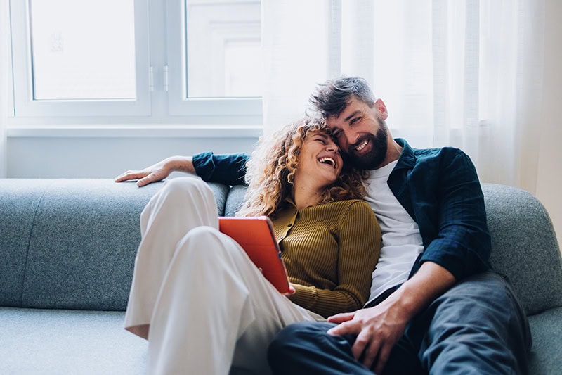 A man and a woman are sitting close together on a grey sofa, laughing and looking happy.