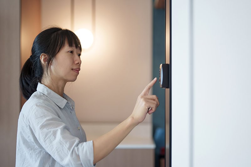 A woman with dark hair tied back in a ponytail is shown from the side as she reaches out to press a button on a small, dark device or digital interface mounted on a white wall.