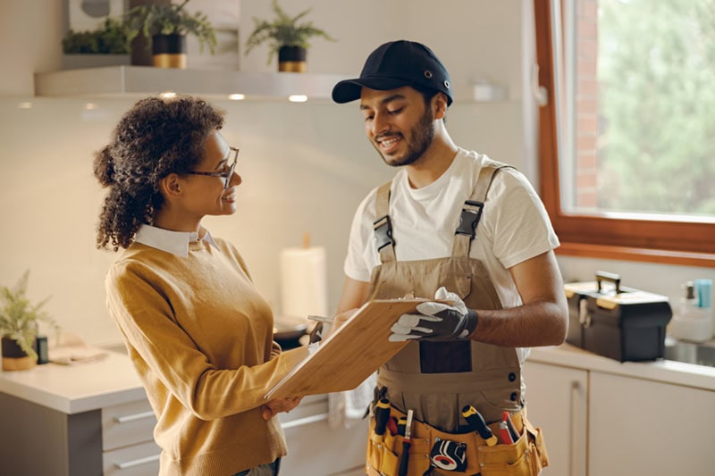 A male service technician and a female homeowner are standing in a kitchen, looking together at a document on a wooden clipboard.