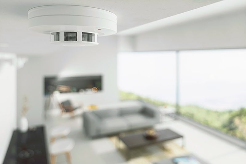 A white smoke detector with a small red indicator light and a central mesh vent is mounted on a white ceiling.