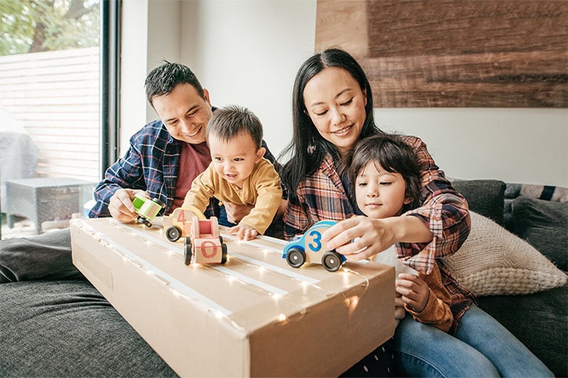 A family of four—a mother, a father, and two young children—are sitting together on a dark couch, playing with toy cars.